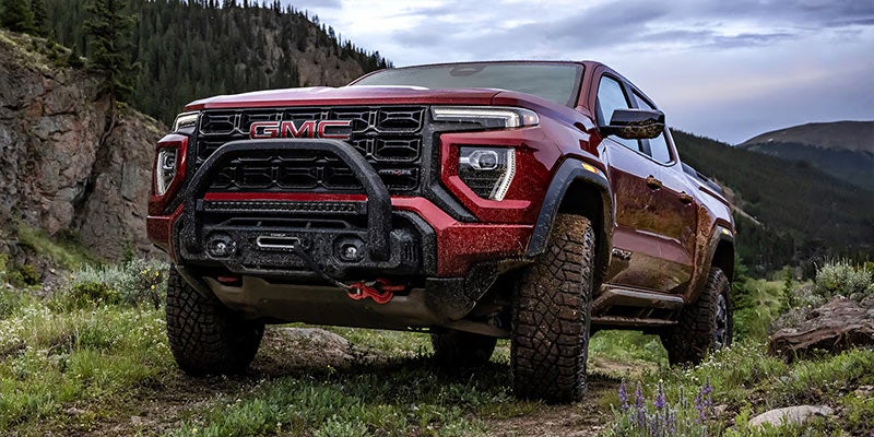 GMC Canyon truck with a winch bumper and off-road tires, parked on a dirt path in a mountainous landscape.