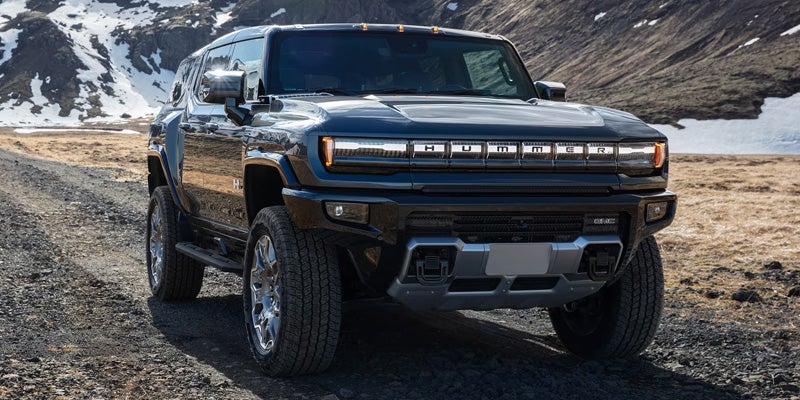 Front view of a GMC Hummer SUV EV, parked on a dirt path with snow-capped mountains and fields in the background.