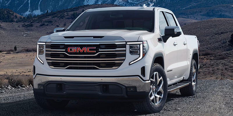 Front view of a GMC Sierra 1500 EV, parked on a dirt path with snow-capped mountains and fields in the background.